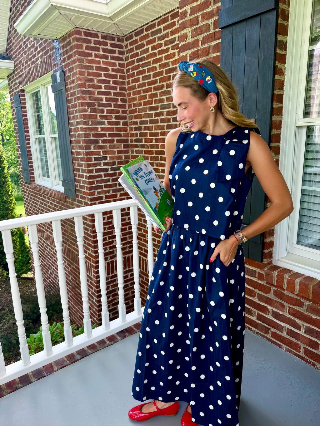 A woman stands on a brick porch wearing a navy sleeveless maxi dress with white polka dots, holding a book. She is wearing a blue patterned headband and red flats.