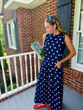 A woman stands on a brick porch wearing a navy sleeveless maxi dress with white polka dots, holding a book. She is wearing a blue patterned headband and red flats.
