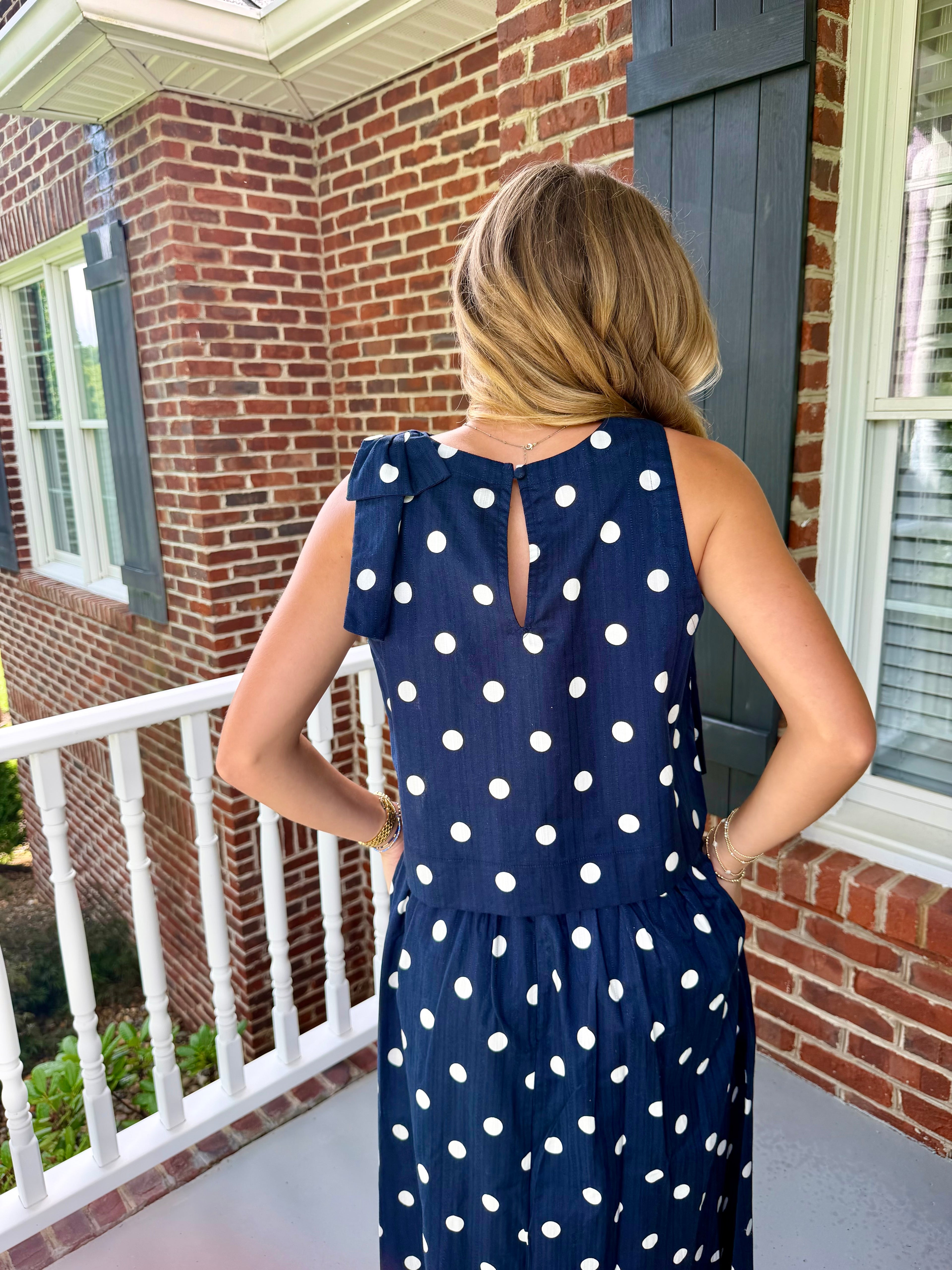 A woman viewed from behind wearing a navy blue sleeveless dress with white polka dots, standing on a brick porch.