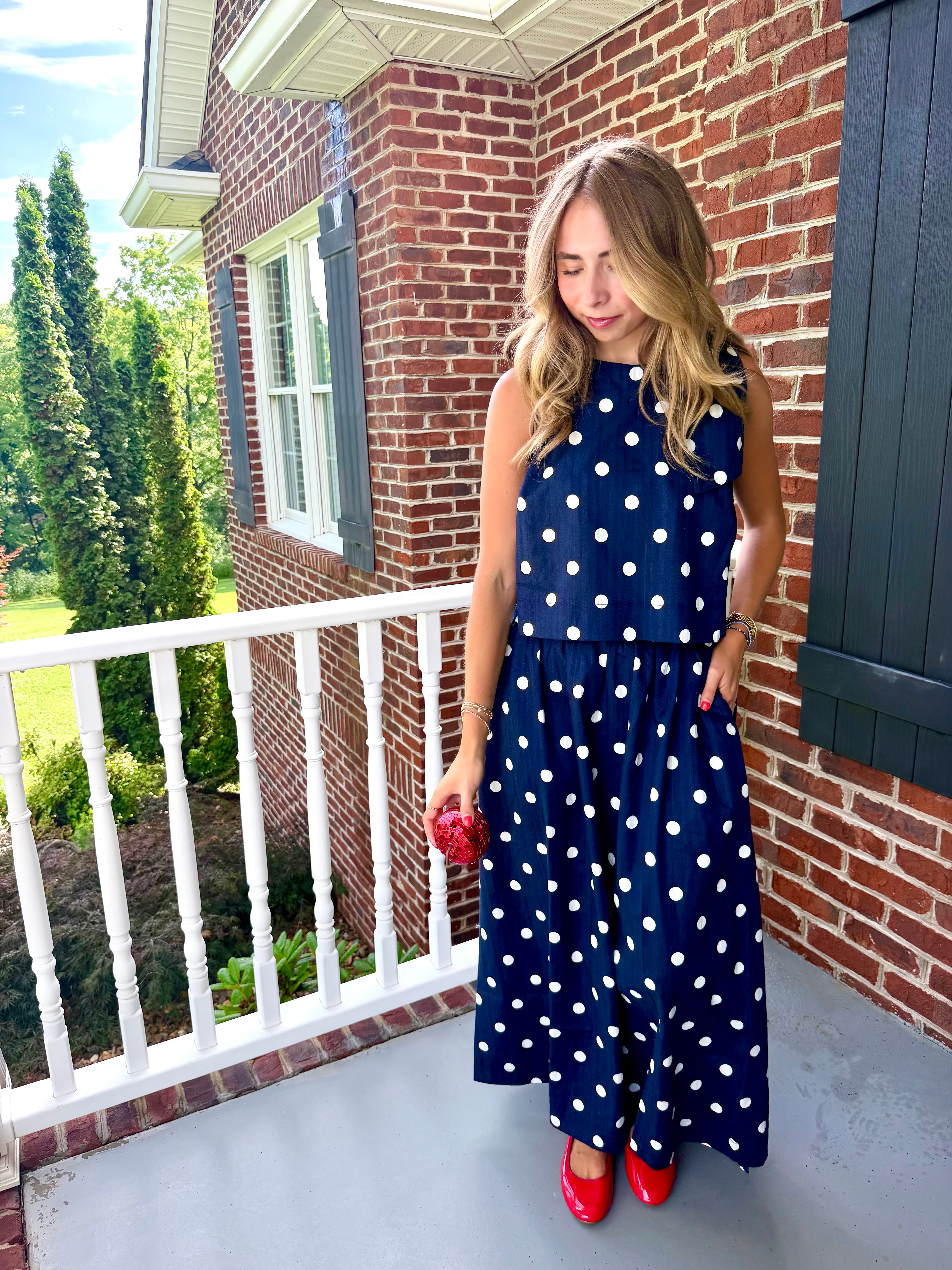 A woman stands on a brick porch wearing a sleeveless navy blue maxi dress with white polka dots, paired with red shoes and holding a small red purse.