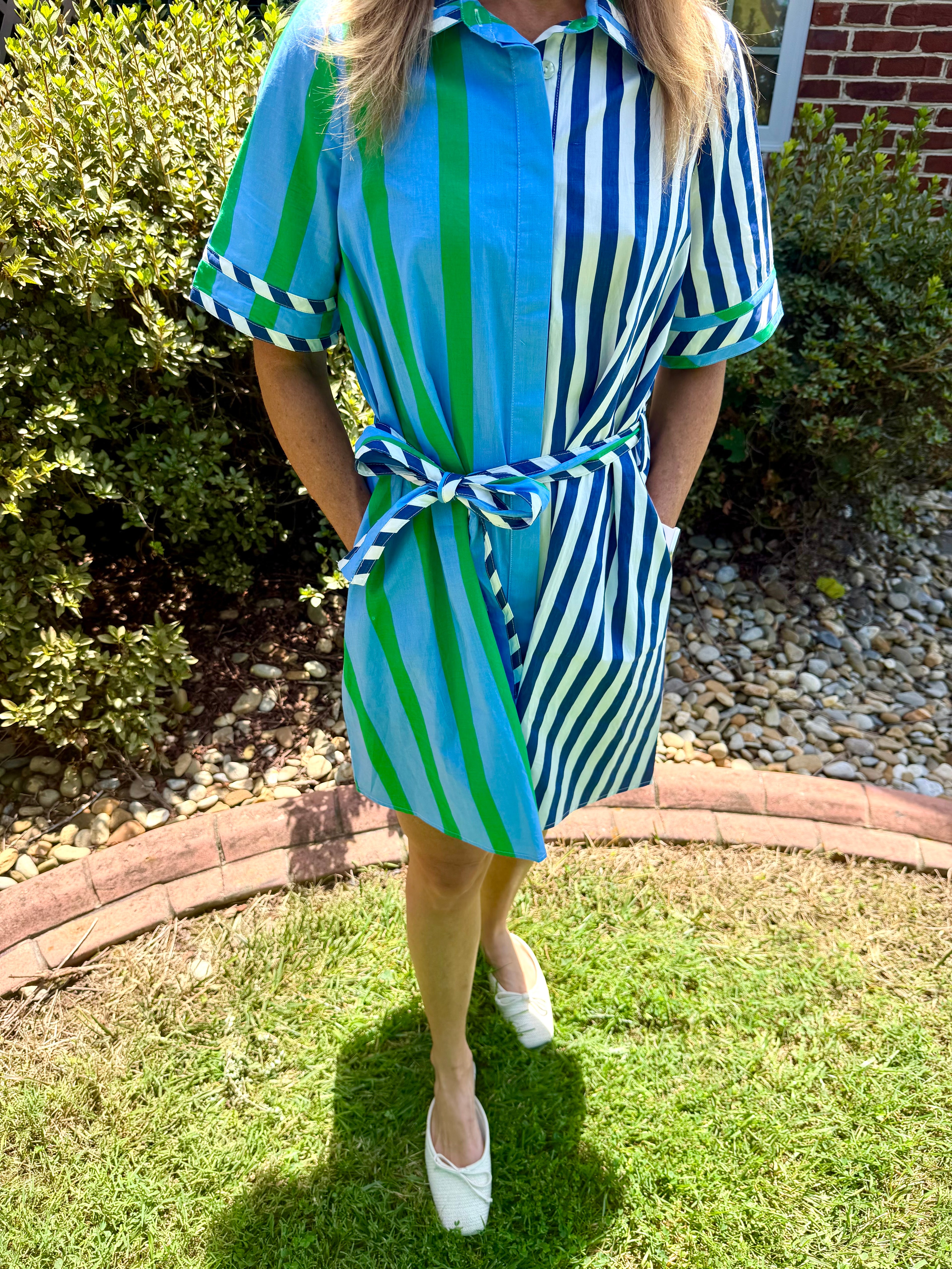 A woman wearing a short-sleeve button-up shirt dress with vertical blue, green, and white stripes, tied at the waist with a matching tie belt, standing on a grassy lawn outdoors.