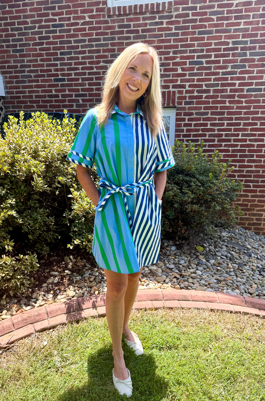 A woman wearing a blue and green striped shirt dress with a tie waist, standing on a grassy lawn in front of a brick house.