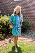 A woman wearing a blue and green striped shirt dress with a tie waist, standing on a grassy lawn in front of a brick house.