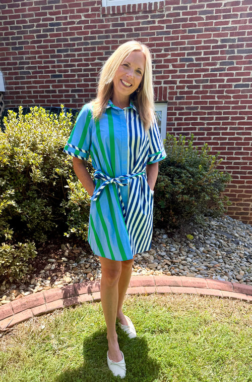 A woman wearing a blue and green striped shirt dress with a tie waist, standing on a grassy lawn in front of a brick house.