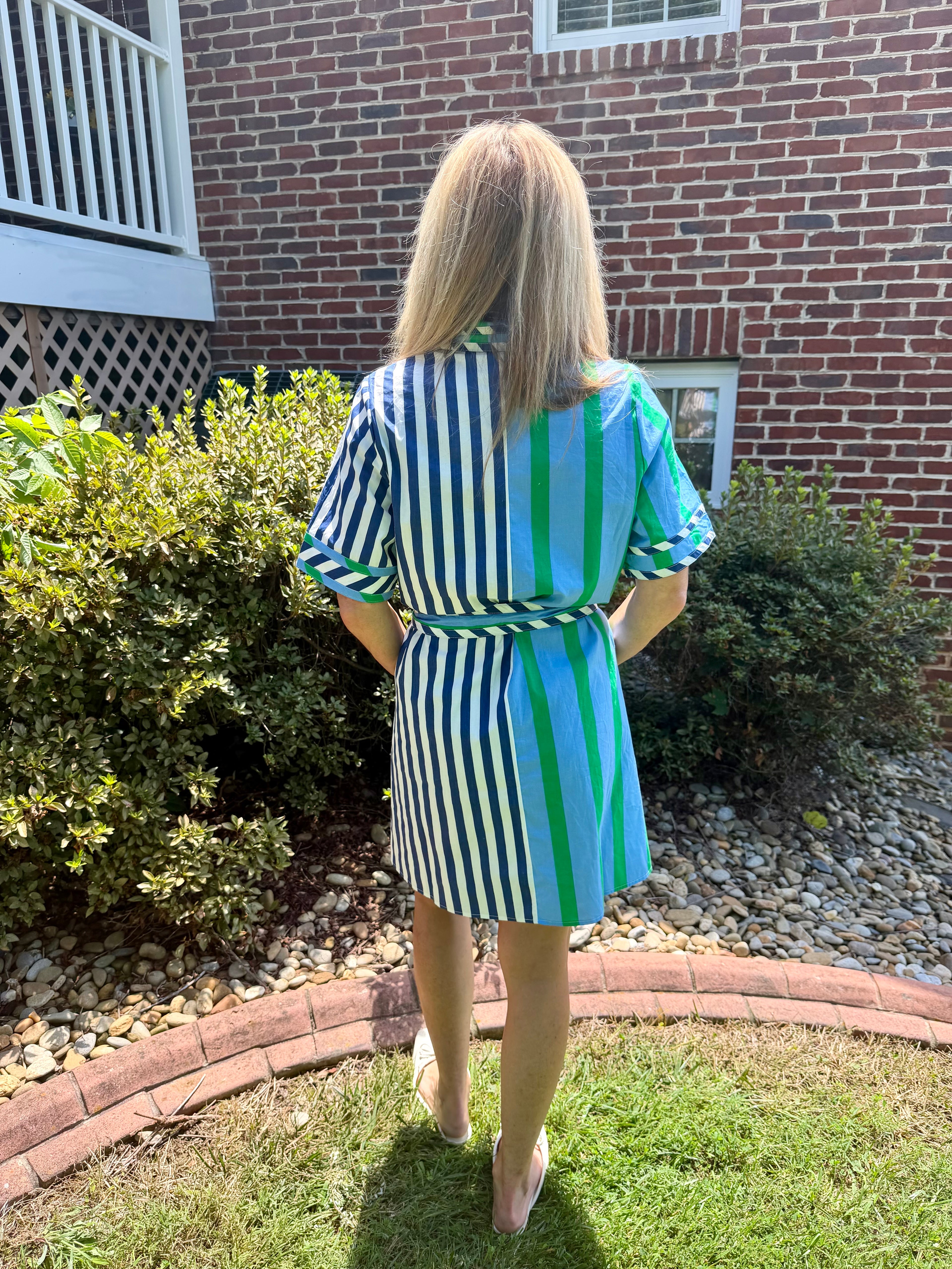 A woman viewed from behind wearing a short-sleeve collared dress with vertical stripes in navy, green, and turquoise, standing on a lawn in front of a brick house.