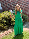 A woman stands outdoors on a grassy lawn in front of a red brick house, wearing a bright green tiered maxi dress with a tie waist. She is holding a small sign that reads 'FIRST DAY OF SCHOOL'.