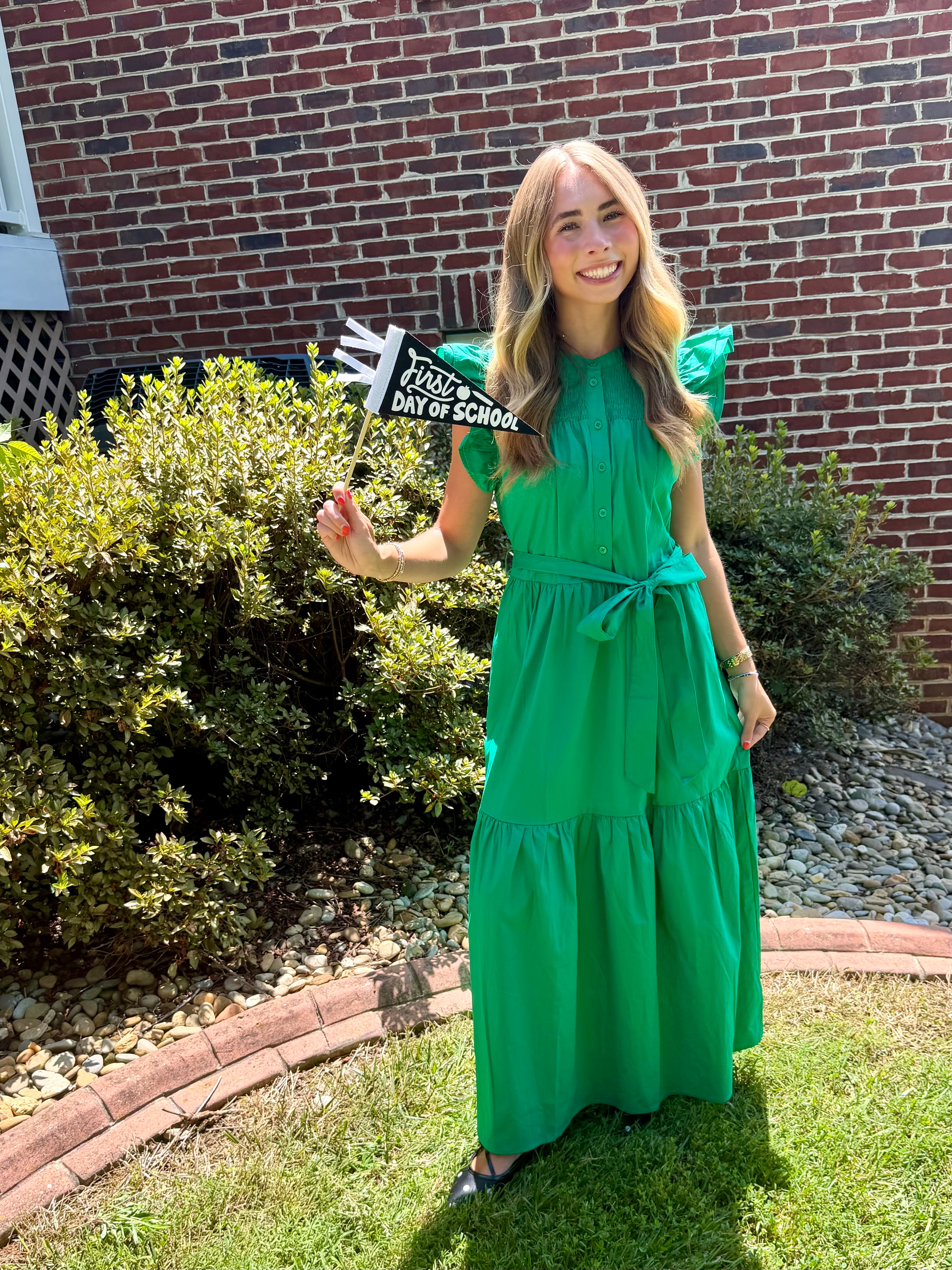 A woman stands outdoors on a grassy lawn in front of a red brick house, wearing a bright green tiered maxi dress with a tie waist. She is holding a small sign that reads 'FIRST DAY OF SCHOOL'.