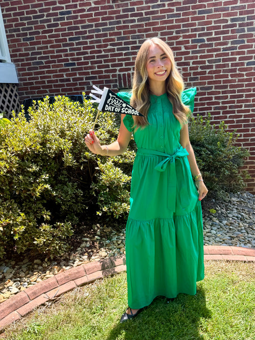 A woman stands outdoors on a grassy lawn in front of a red brick house, wearing a bright green tiered maxi dress with a tie waist. She is holding a small sign that reads 'FIRST DAY OF SCHOOL'.