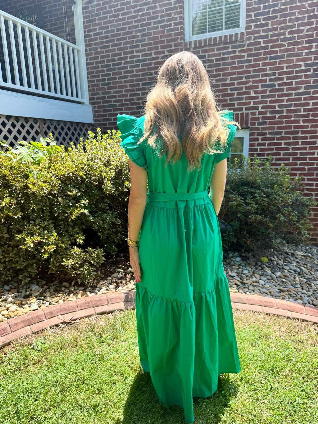 A woman viewed from behind wearing a bright green tiered midi dress with ruffled short sleeves and a matching belt, standing on a grassy lawn in front of a brick house.