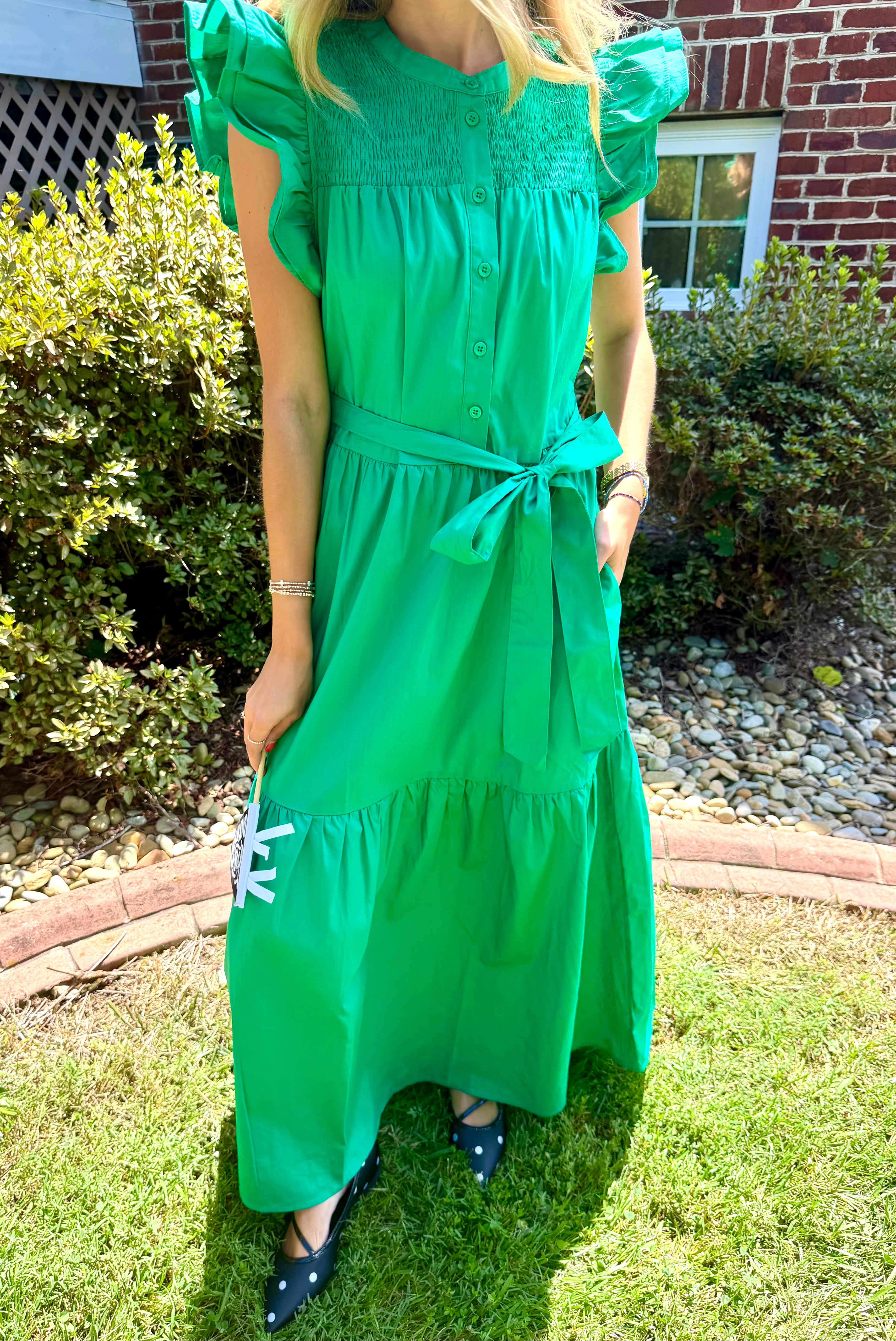 A woman wearing a bright green midi dress with puff sleeves, a smocked bodice, and a matching waist tie, standing outdoors on a grassy lawn in front of a brick house.
