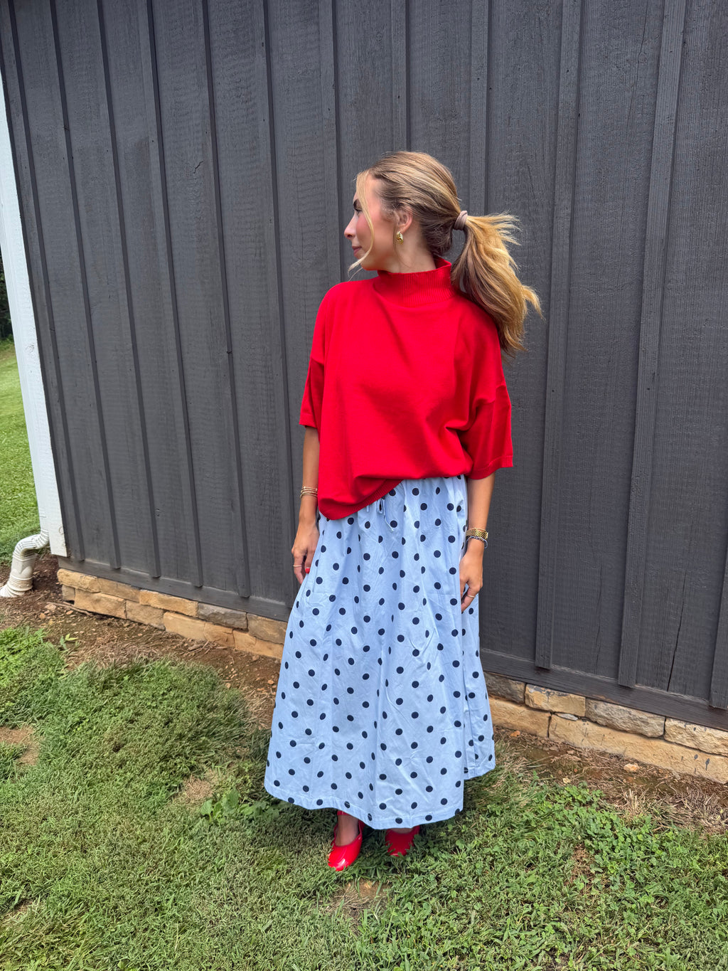 A woman stands outdoors in front of a dark gray wooden fence, wearing a bright red mock-neck top and a light blue midi skirt with black polka dots, paired with red shoes.