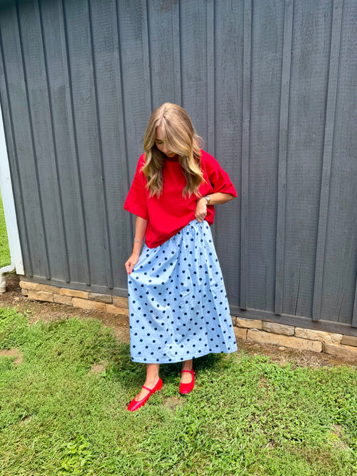 A woman stands outdoors in front of a dark gray wooden fence on a grassy lawn, wearing a bright red short-sleeve top, a light blue midi skirt with black polka dots, and red flat shoes.