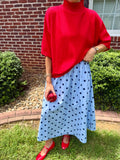A person outdoors wearing a bright red turtleneck sweater and a light blue midi skirt with dark polka dots, paired with red shoes and holding a red apple.