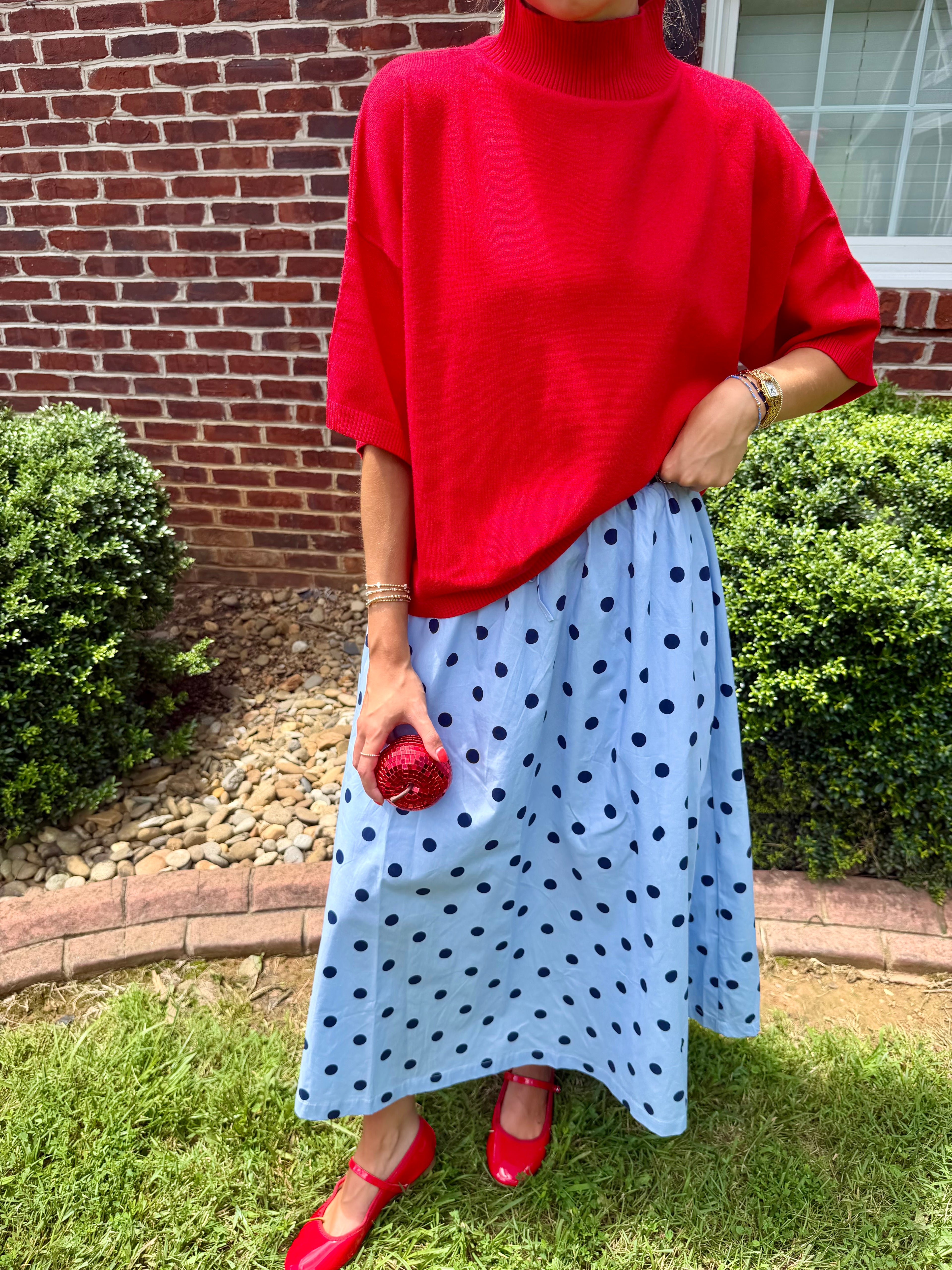 A person outdoors wearing a bright red turtleneck sweater and a light blue midi skirt with dark polka dots, paired with red shoes and holding a red apple.