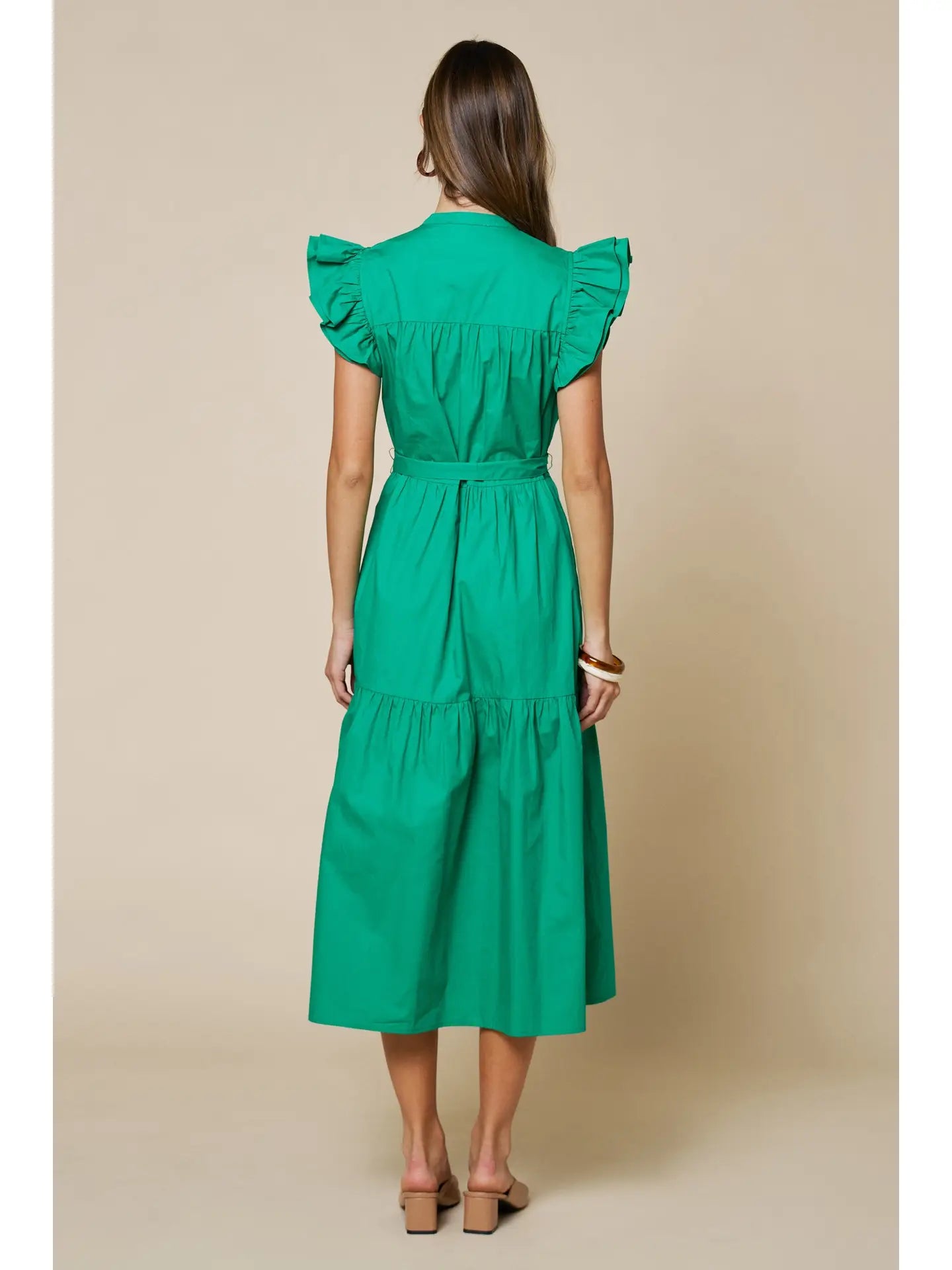 A woman viewed from the back wearing a bright green midi dress with ruffled cap sleeves and a tie waist, standing against a plain beige studio background.