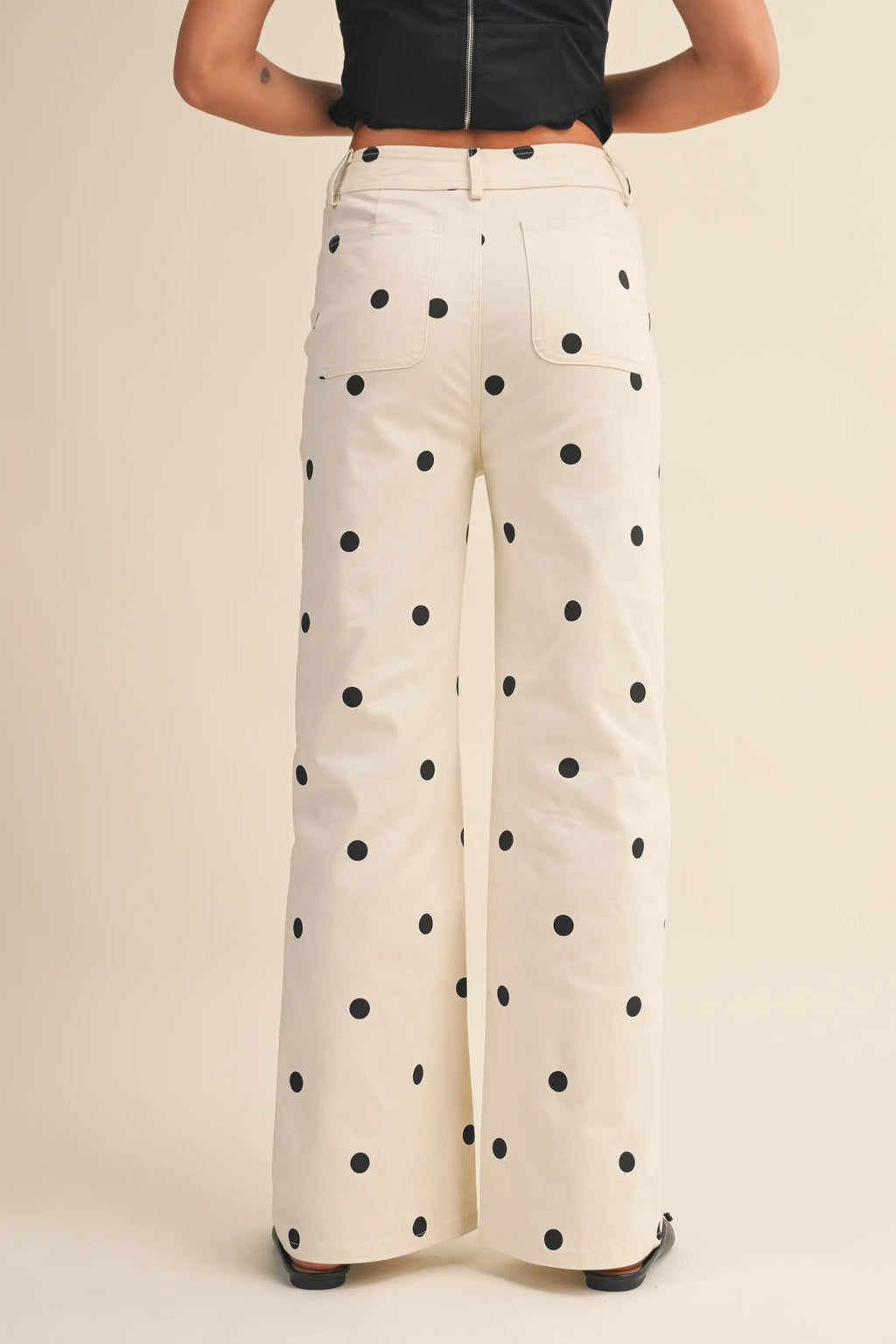 A person shown from the back wearing high-waisted, wide-leg pants in an off-white fabric with a black polka dot pattern, paired with a black top and black shoes, against a solid beige studio background.