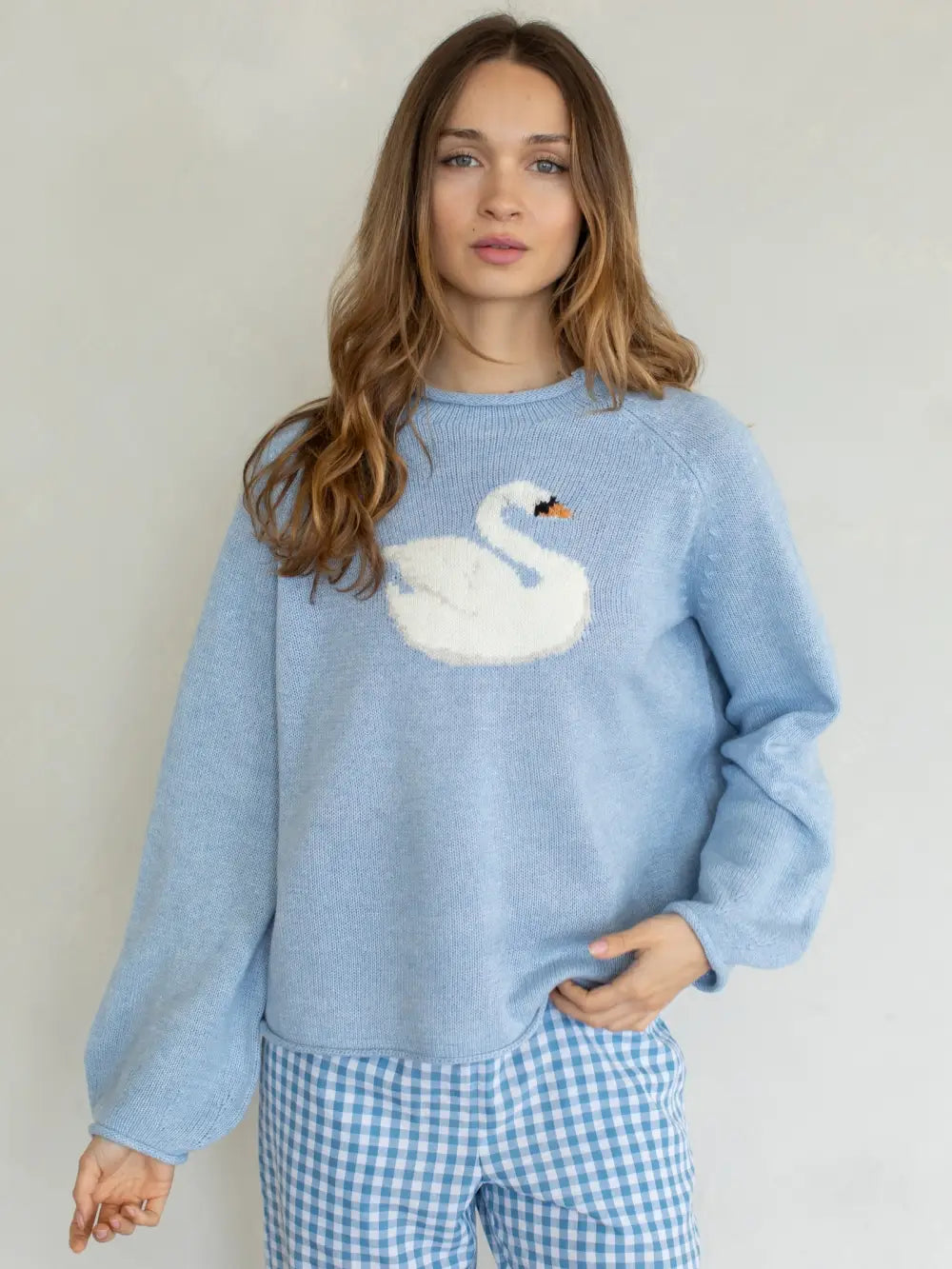 A woman wearing a light blue crewneck sweater with a white swan motif on the chest and blue-and-white gingham pants, standing against a plain light gray wall.