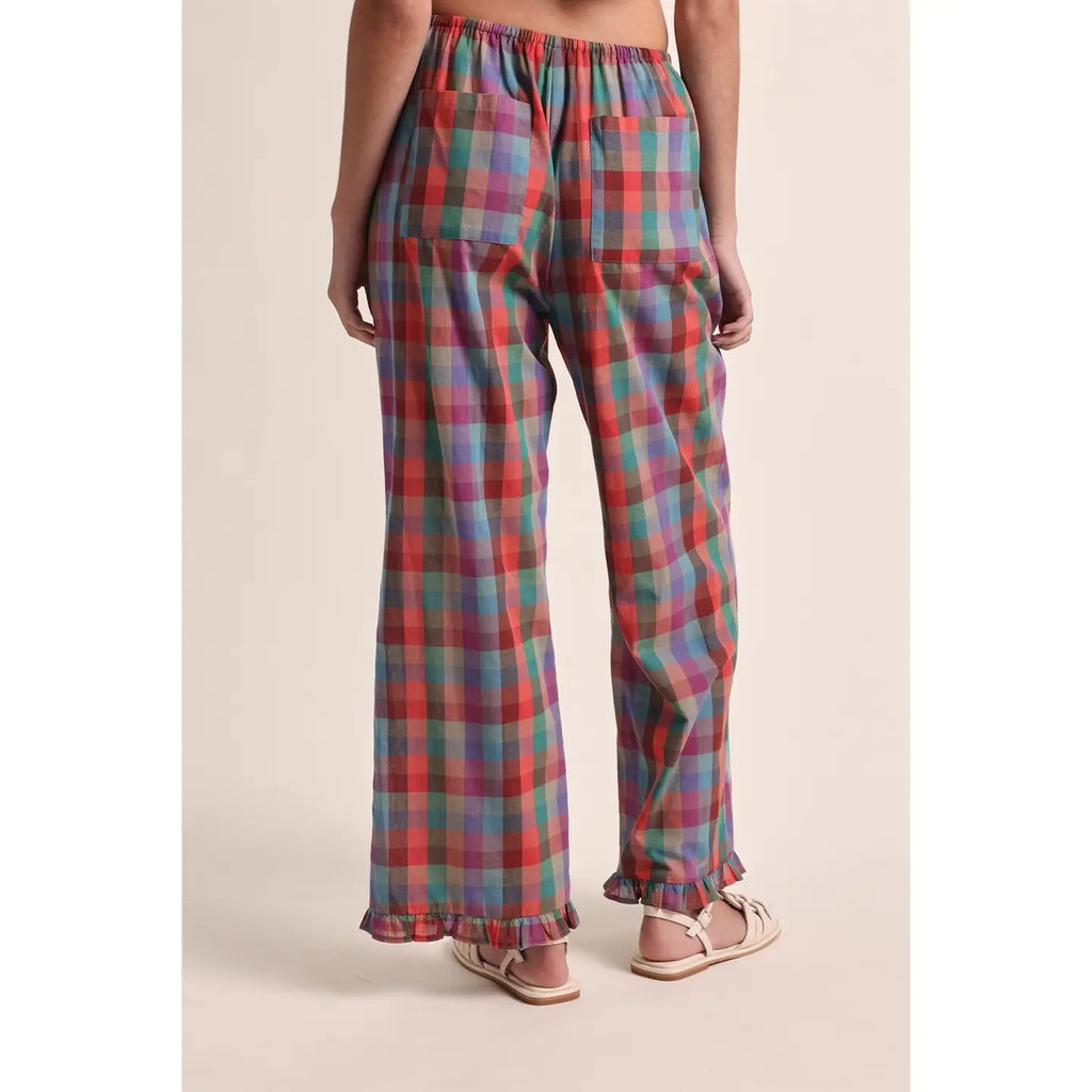 A person shown from the back wearing loose plaid pants with an elastic waistband and ruffled hems, paired with light pink sandals, against a plain beige studio background.