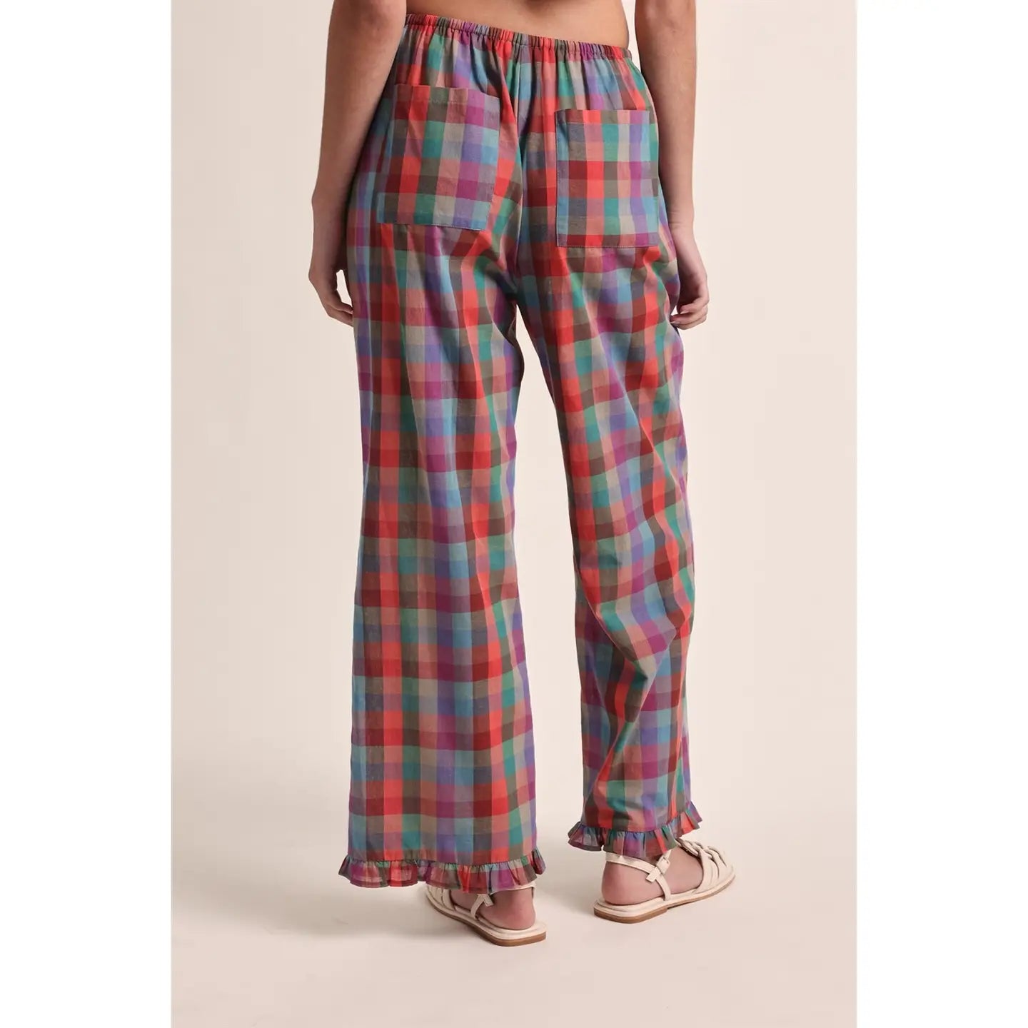 A person shown from the back wearing loose plaid pants with an elastic waistband and ruffled hems, paired with light pink sandals, against a plain beige studio background.