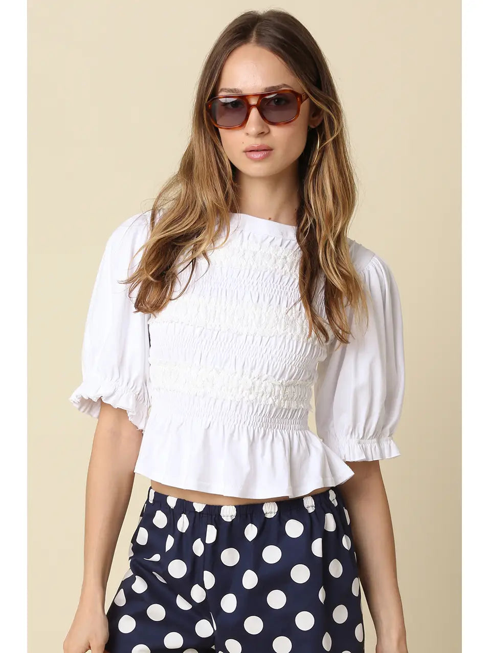 A woman modeling a white short-sleeve crop top with puff sleeves and a smocked bodice, paired with navy blue and white polka dot pants, against a solid beige background.