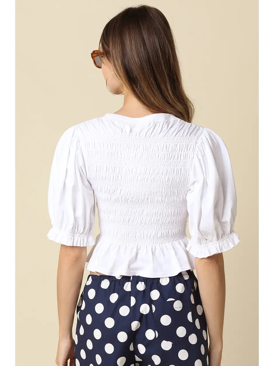 A woman shown from the back wearing a white smocked puff-sleeve top and navy blue pants with white polka dots, set against a solid beige background.