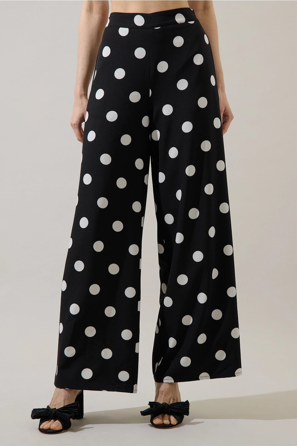 Black wide-leg pants with a white polka dot pattern, worn by a model with black bow sandals against a neutral studio background.