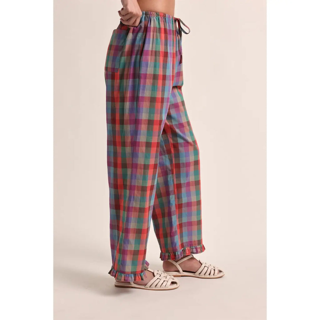 A person wearing loose, wide-leg plaid pants in a multicolor pattern of red, green, blue, and purple, paired with white strappy flat sandals, shown from the waist down against a solid beige background.