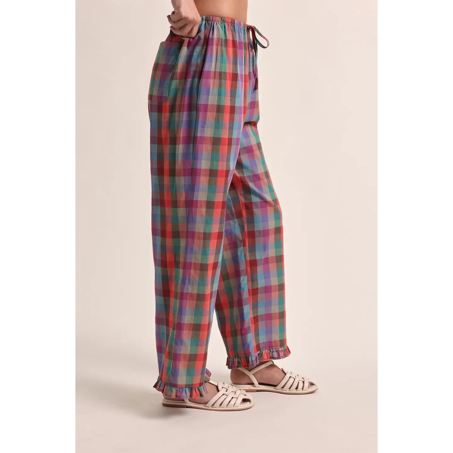 A person wearing loose, wide-leg plaid pants in a multicolor pattern of red, green, blue, and purple, paired with white strappy flat sandals, shown from the waist down against a solid beige background.