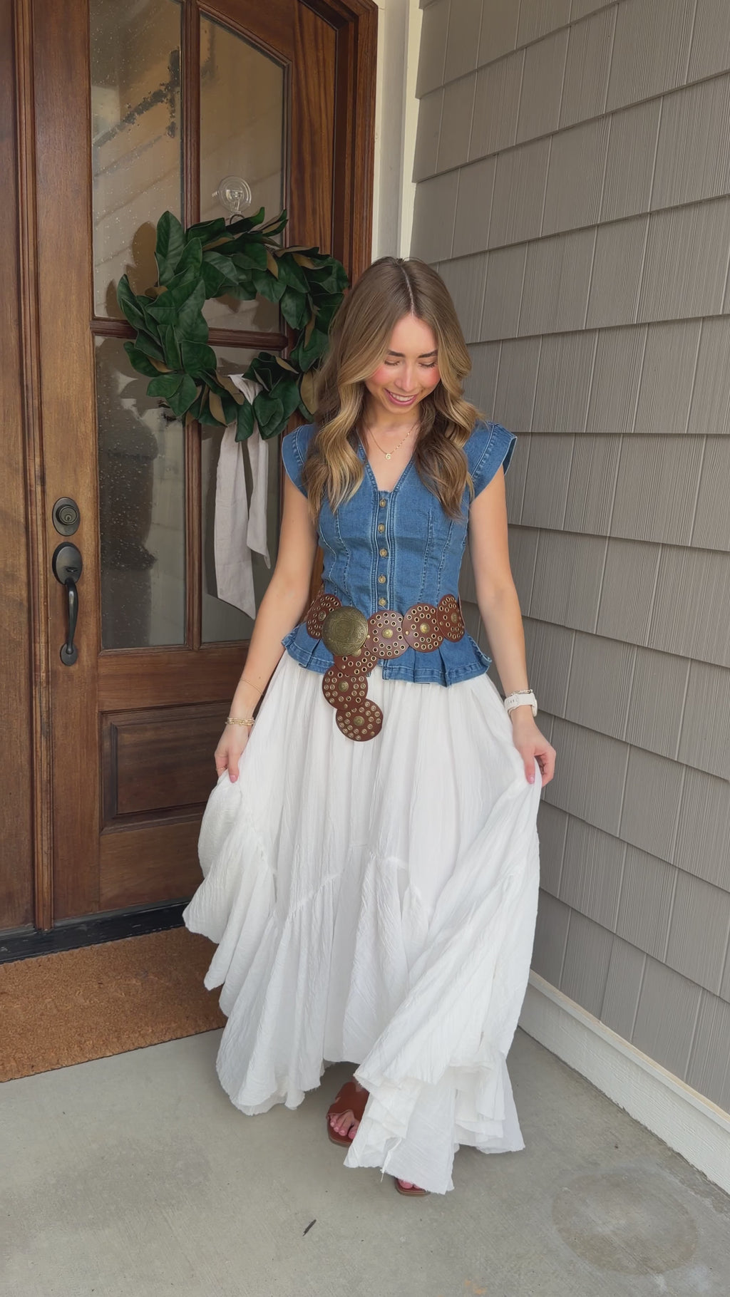Woman in a blue top and white skirt standing outside a house.