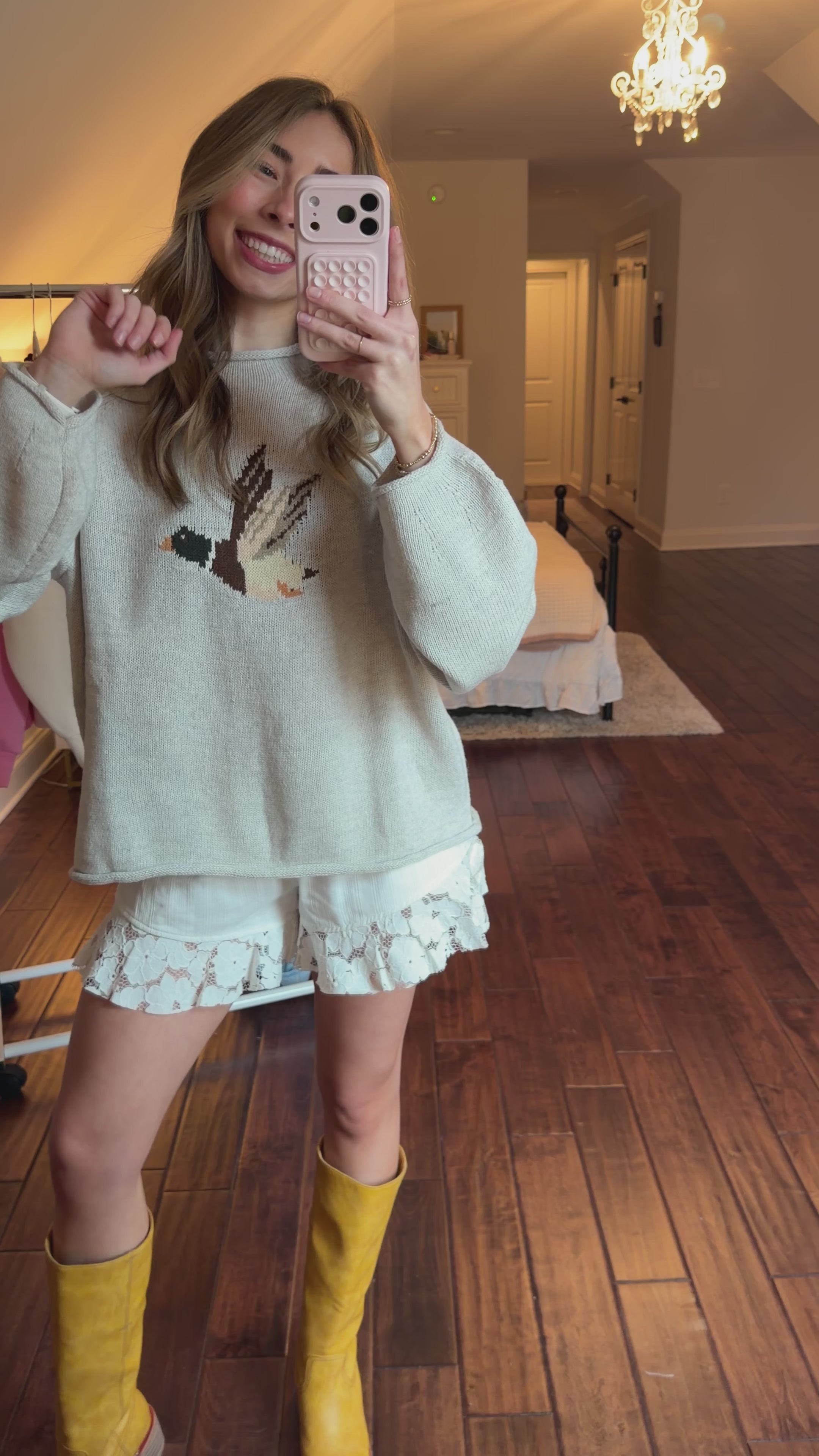Women taking a mirror selfie in a room with wooden flooring and a clothing rack. Women is wearing a duck sweater, white shorts, and yellow boots. 