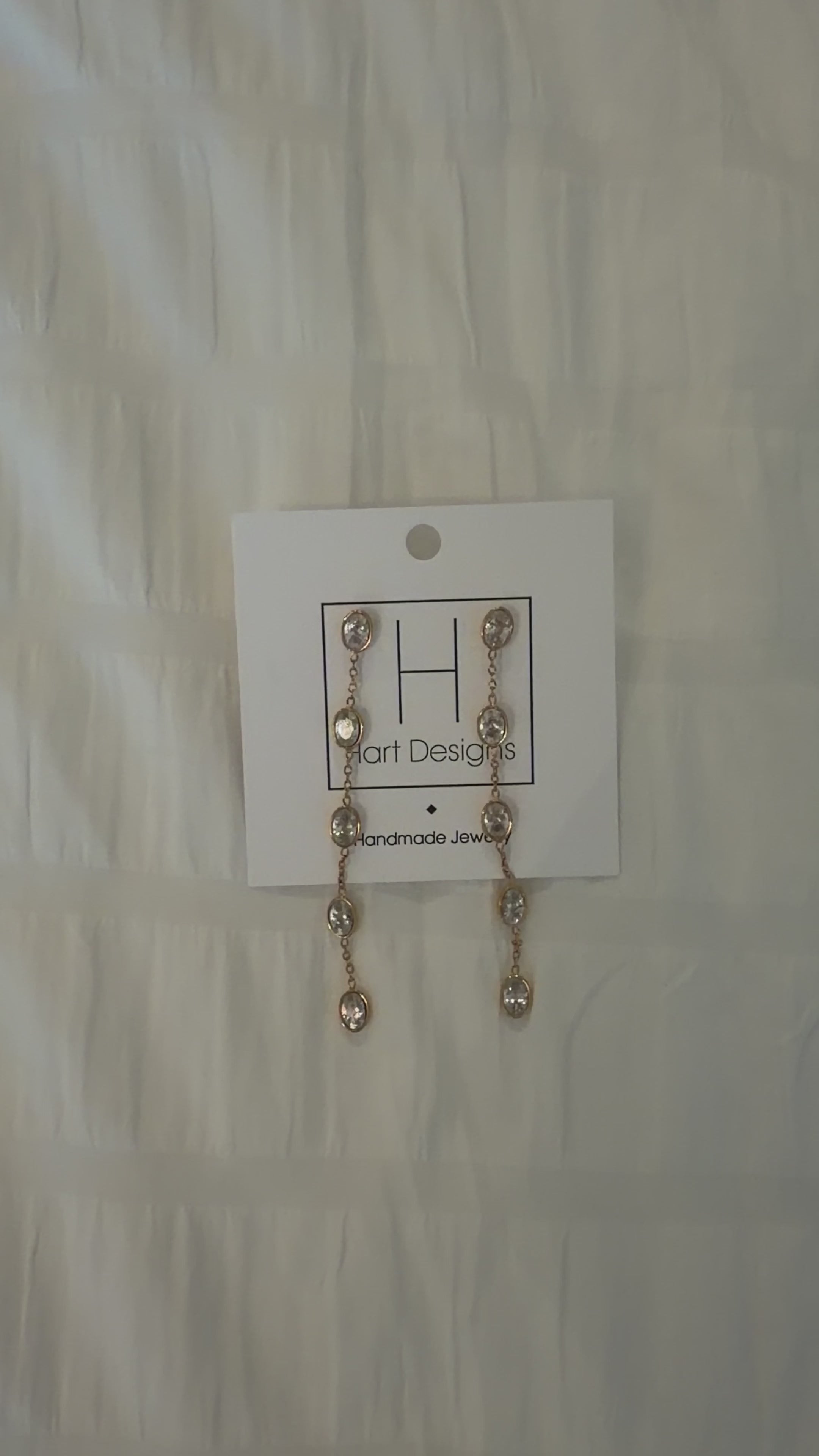 Gold earrings with clear stones on a white card with 'Hart Designs' branding, placed on a textured beige surface.
